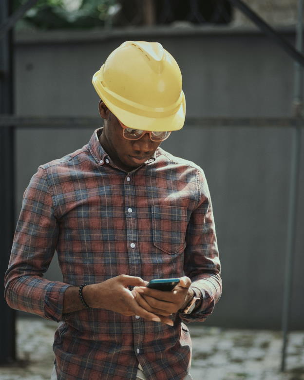 Man met bril en gele helm staat op werf met smartphone in de hand